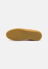 Gummi-sohliger Schuh mit einem strukturierten orangen Obermaterial und einer gemusterten Gummisohle, die in der Mitte ein kreisförmiges Detail aufweist.