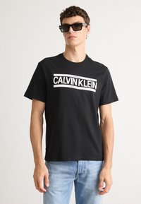 Black cotton t-shirt with white “CALVIN KLEIN” logo, short sleeves, crew neck, paired with blue denim jeans and dark rectangular sunglasses.