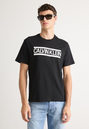 Black cotton t-shirt with white “CALVIN KLEIN” logo, short sleeves, crew neck, paired with blue denim jeans and dark rectangular sunglasses.