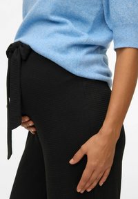 Svetlo modra pletena majica s kratkimi rokavi, kombinirana z teksturiranimi črnimi hlačami z detajlom zavezovanja v pasu. Roka počiva na hlačah.