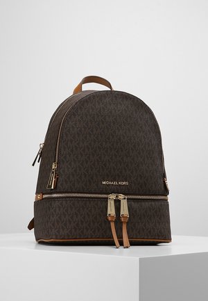 Sac à dos marron Michael Kors avec fermetures éclair dorées, motif logo MK et tirettes en cuir, posé sur un socle blanc sur fond uni.