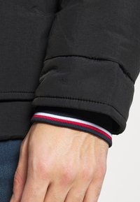 Manica di una giacca a vento nera in tessuto strutturato. Il polsino presenta una fascia a coste con strisce rosse, bianche e blu. Una mano è visibile sulla manica.