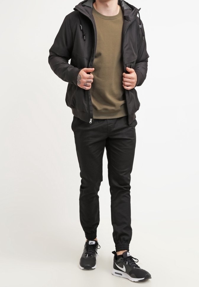 Veste noire à fermeture éclair avec capuche, t-shirt vert à col rond, pantalon noir à taille élastique et baskets noires Nike avec accents blancs.