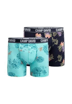 Lächelnder muskulöser Mann mit kurzen braunen Haaren, der florale Boxer mit "CAMP DAVID"-Bund vor einfarbigem Hintergrund trägt.