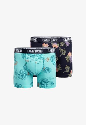 Smiling muscular man with short brown hair wearing floral patterned boxer briefs with "CAMP DAVID" waistband against plain background.