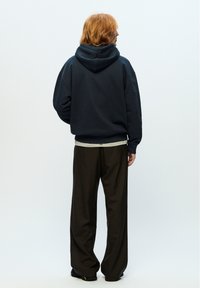 Navy hoodie made of soft fabric, relaxed fit, with a kangaroo pocket. Paired with loose brown trousers and black shoes.