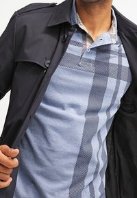 Veste marine foncée sur une chemise bleu clair avec un motif à carreaux et rayures, présentant une texture lisse et un col boutonné.