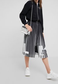 Schwarze Bomberjacke mit Reißverschluss und weißem Logo, kombiniert mit einem schwarz-weiß gestreiften Midirock. Silberne Handtasche mit Krokoprägung und weiße Turnschuhe.