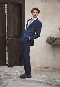 Homme en costume bleu marine et cravate rayée, se penchant en arrière, les mains dans les poches, près d'une porte en bois ouverte et d'une fontaine en pierre.