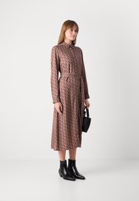 Donna che indossa un vestito midi a maniche lunghe con motivo, una cintura, stivali neri alla caviglia e tiene in mano una piccola borsa nera, in piedi su uno sfondo bianco.