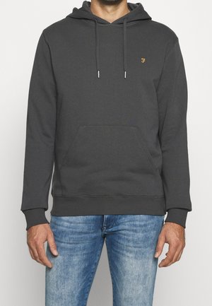 Mężczyzna ubrany w ciemnoszarą bluzę z kapturem, z przednią kieszenią i małym żółtym logo, w połączeniu z wyblakłymi niebieskimi dżinsami.