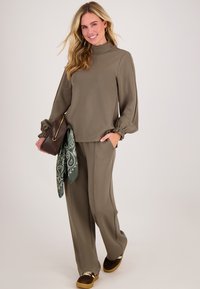 Vrouw met lang blond haar, gekleed in een olijfgroene top met hoge hals en broek, die een bruine clutch met een gebloemde sjaal vasthoudt en glimlacht.