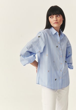 Kobieta o ciemnych włosach, ubrana w jasnoniebieską, prążkowaną koszulę na guziki z drobnym kwiatowym i pszczelim haftem, w połączeniu z białymi spodniami, patrząca na bok.