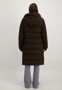 Brun pufferjacka med huva, med ett kviltat mönster och långa ärmar. Jackan går ner till mitten av låret och är parat med ljusblå utsvängda jeans.