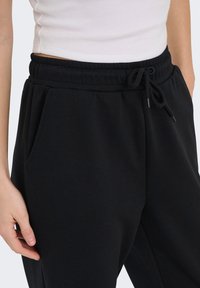 Κοντινό πλάνο ενός ατόμου που φοράει μαύρο φούτερ παντελόνι με ελαστική μέση και κορδόνι, συνδυασμένο με λευκό ριγωτό κορμάκι cropped.