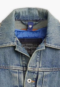 Jachetă din denim albastru deschis cu guler clasic, cusături din cupru și căptușeală de un navy. Prezintă un etichetă de marcă și închidere cu butoane snap.