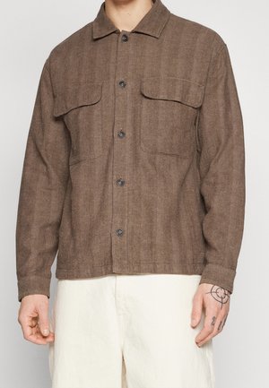 Homme portant une veste marron à motif chevrons avec deux poches poitrine et un pantalon écru, montrant sa main droite tatouée.