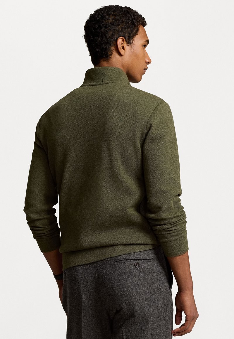 Olivgrüner Strickpullover mit Stehkragen, gerippten Bündchen und strukturiertem Finish, kombiniert mit grauen Hosen, die eine Gesäßtasche haben.