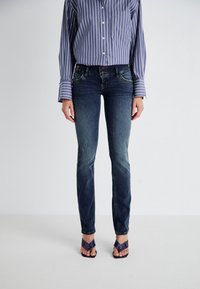 Jeans en denim bleu foncé avec une coupe slim, un léger délavage et des détails cousus. Associés à une chemise à rayures avec des boutons et des tongs violettes.