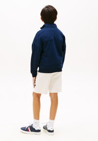 Μπλε ναυτικό φούτερ με ανεβασμένο γιακά, άνετη εφαρμογή· συνδυασμένο με λευκά σορτσάκια. Φορεμένο με μπλε ναυτικά αθλητικά παπούτσια με κόκκινες και λευκές λεπτομέρειες.