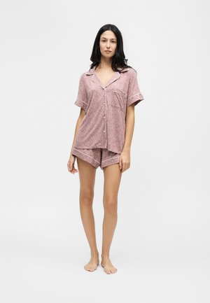Femme aux longs cheveux foncés portant un ensemble de pyjama rose à manches courtes avec un liseré blanc, debout pieds nus sur un fond uni.