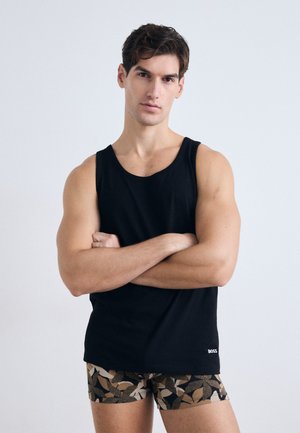 Canottiera nera senza maniche in cotone. Il modello indossa pantaloni boxer beige e marrone con disegni di foglie. Branding semplice sulla maglietta.