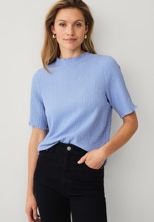 Kvinde med skulderlangt blondt hår iført sølvfarvede øreringe, lyseblå struktureret kortærmet top og sorte bukser med høj talje.