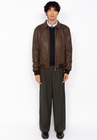 Chaqueta de cuero marrón con cuello tipo camisa, cierre de cremallera y puños acanalados, llevada sobre un suéter oscuro, combinada con unos pantalones grises anchos.