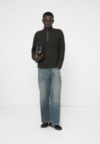 Dark green ribbed sweater with a quarter-zip, baggy light blue jeans, and black slip-on shoes, holding a black bag. Minimalist background.