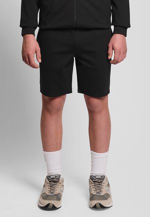 Person som bär svarta shorts, svart zip-up jacka, vita strumpor och beige New Balance 991-sneakers som står på ett grått golv.