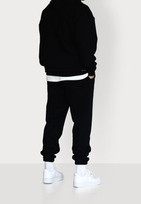 Persona vestida con sudadera negra y pantalones de chándal negros, con calcetines blancos y zapatillas blancas, de pie con las manos en los bolsillos contra un fondo blanco.