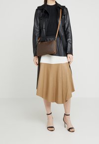 Černý kožený trenčkot přes černý pletený top, doplněný hnědou skládací sukní. Hnědá kabelka se vzorem a béžovým popruhem. Černé sandály na podpatku.