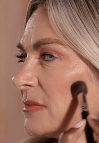 Aplicación de maquillaje sobre una mejilla lisa y de piel clara, con una textura natural. Se utiliza un pincel de cerdas oscuras para iluminar.