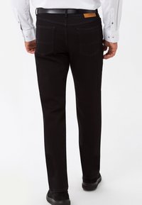 Hombre vestido con pantalones negros de pierna recta, zapatos negros y una camisa blanca de manga larga, de pie mirando hacia atrás sobre un fondo blanco.