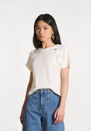 Jeune femme portant un haut crème à manches courtes avec des boutons dorés sur les manches et un jean bleu taille haute sur fond blanc.