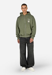 Zöld zipzáras kapucnis felső elülső zsebekkel, szürke farmernadrág és fehér sportcipő. A kapucnis felső bal melle részén logó részlet található. Kényelmes illeszkedés.