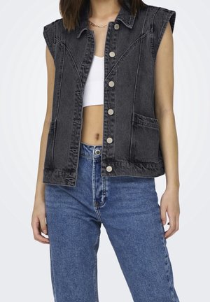 Mouwloos zwart denimvest met zilveren knopen over een witte crop top, gecombineerd met een high-waisted blauwe spijkerbroek bij een staand persoon.