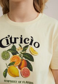 Kremo spalvos marškinėliai su citrusinių vaisių grafiniu dizainu, vaizduojančiu apelsinus, citrinas ir laimus, su juodais užrašais "Citrico."