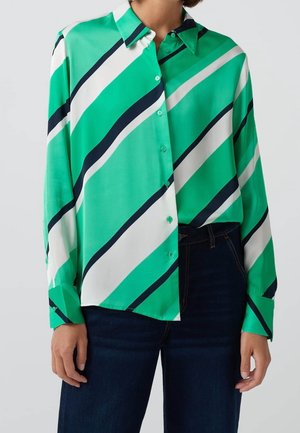 Mujer que lleva una camisa abotonada de manga larga con rayas diagonales verdes, blancas y azul marino, metida dentro de unos vaqueros azul oscuro.