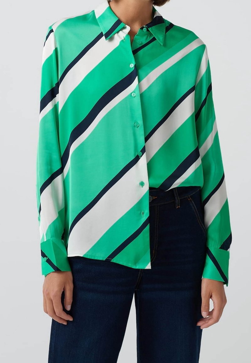 Femme portant une chemise boutonnée à manches longues à rayures diagonales vertes, blanches et bleu marine, rentrée dans un jean bleu foncé.