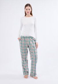 Kobieta stoi boso, ubrana w białą bluzkę z długim rękawem i luźne zielone spodnie w kratę, z rękami w kieszeniach, na białym tle.