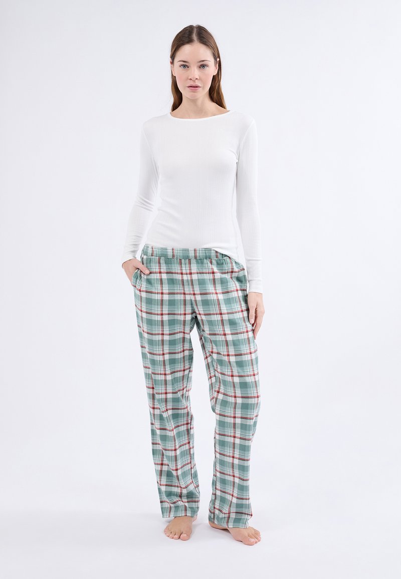 Kobieta stoi boso, ubrana w białą bluzkę z długim rękawem i luźne zielone spodnie w kratę, z rękami w kieszeniach, na białym tle.