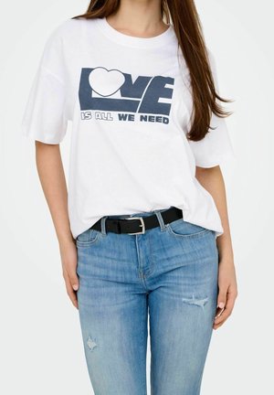 Femme portant un t-shirt blanc avec le texte "LOVE is all we need", assorti à un jean bleu clair déchiré et une ceinture noire, sur un fond uni.