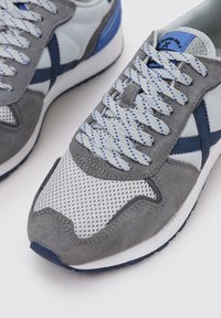 Zapatillas gris y azul con parte superior de malla y ante, cordones redondos, cuello acolchado y una entresuela blanca, que presenta un diseño de cordones estampado.