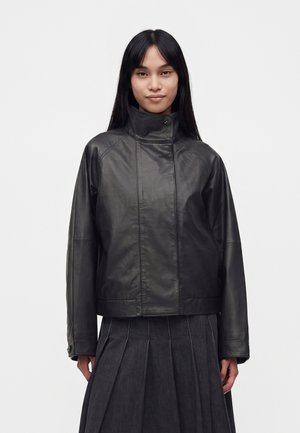 Jeune femme aux longs cheveux noirs portant une veste en cuir noire à col montant et une jupe plissée foncée sur un fond uni.