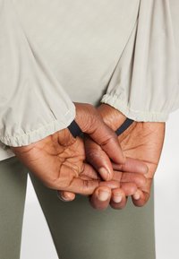 Manos de piel oscura sosteniendo bucles negros en el pulgar, unidos a unas mangas largas beige, y usando leggings verdes.