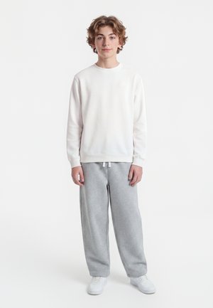 Adolescent aux cheveux bouclés portant un t-shirt blanc à manches longues, un pantalon de survêtement gris et des baskets blanches, debout devant un fond blanc.