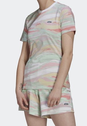 Osoba ubrana w pastelowy, tie-dye t-shirt Adidas oraz pasujące szorty z naszywkami z logo, stojąca z rękami swobodnie opuszczonymi wzdłuż ciała.