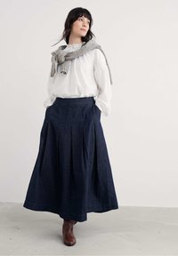 Frau trägt eine weiße Bluse, einen dunkelblauen, plissierten Midirock, braune Stiefel und einen grauen Pullover, der über ihren Schultern drapiert ist, und steht auf einem grauen Boden.