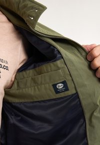 Interiorul jachetei de culoare verde măsliniu prezintă o căptușeală albastră, un buzunar mic și un eticheta cu textul "Pivetro Industries". Se observă închiderea cu fermoar.